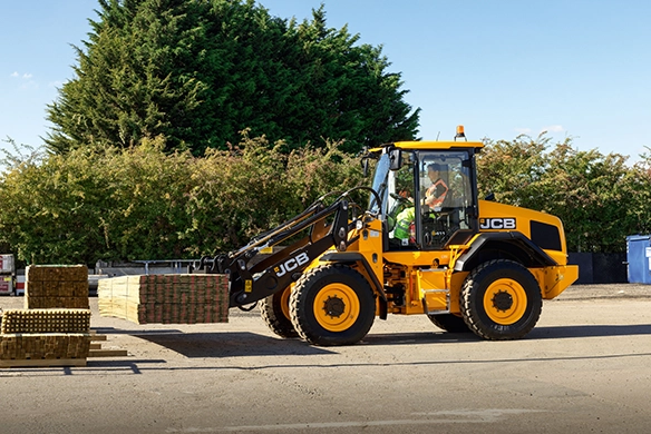 JCB 411 wheel loader transporting palletized materials at an outdoor construction site with trees in background