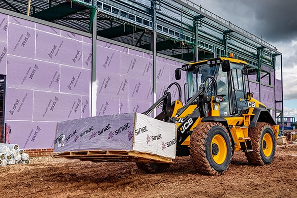 JCB 411 wheel loader carrying pink insulation boards at a construction site with steel framework and a cloudy sky