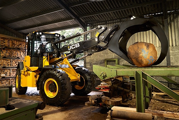 JCB 417 wheel loader with log grapple attachment handling a large log in an indoor sawmill facility with stacked lumber in background