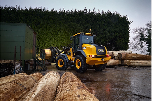 JCB 417 wheel loader positioned near large logs at an outdoor timber processing yard with wood chipper equipment visible