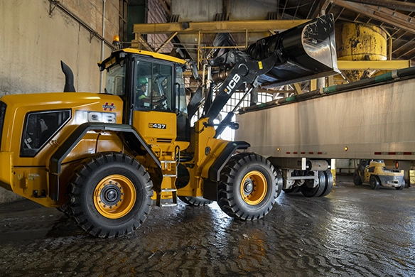 JCB 437 wheel loader lifting bucket to load material into a truck inside an industrial facility