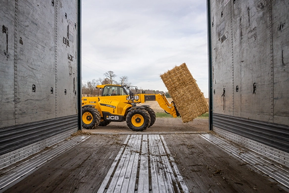 JCB 542-70 telescopic handler loading a rectangular straw bale inside a curtain-side trailer