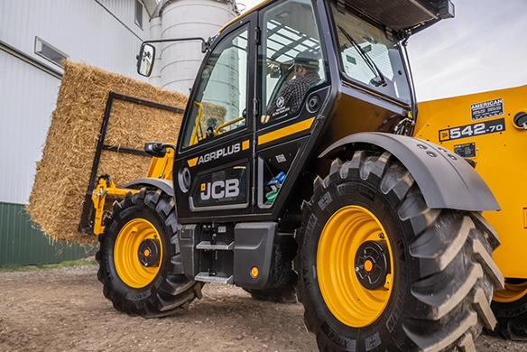 Low-angle view of a JCB 542-70 telescopic handler carrying stacked straw bales beside a farm building