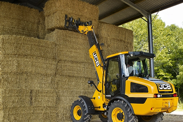 Rear three-quarter view of JCB TM220 telescopic wheel loader lifting straw bales in a farm shed