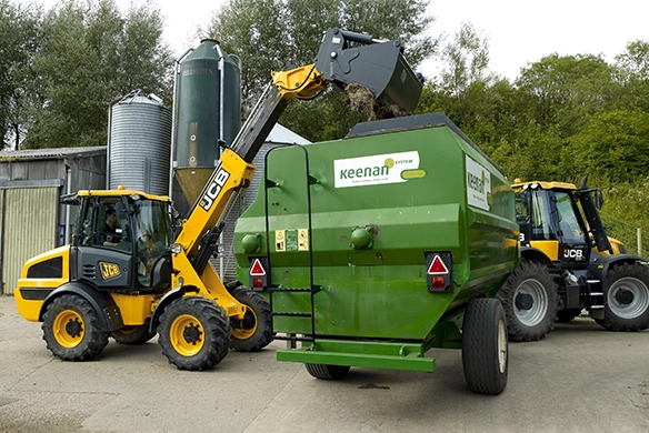 JCB TM220 telescopic wheel loader feeding material into a mixer wagon in an agricultural yard
