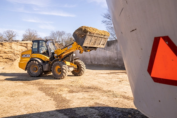 JCB TM320 telescopic wheel loader carrying a full bucket of organic material in a farm yard